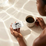 A person holds a Lea Gogo lactase metal tin in their hand next to a cup of black coffee on a sunlit cream surface.